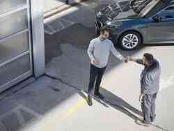 Zwei Männer unterhalten sich auf einem Parkplatz. Hinter ihnen steht ein graues Fahrzeug.